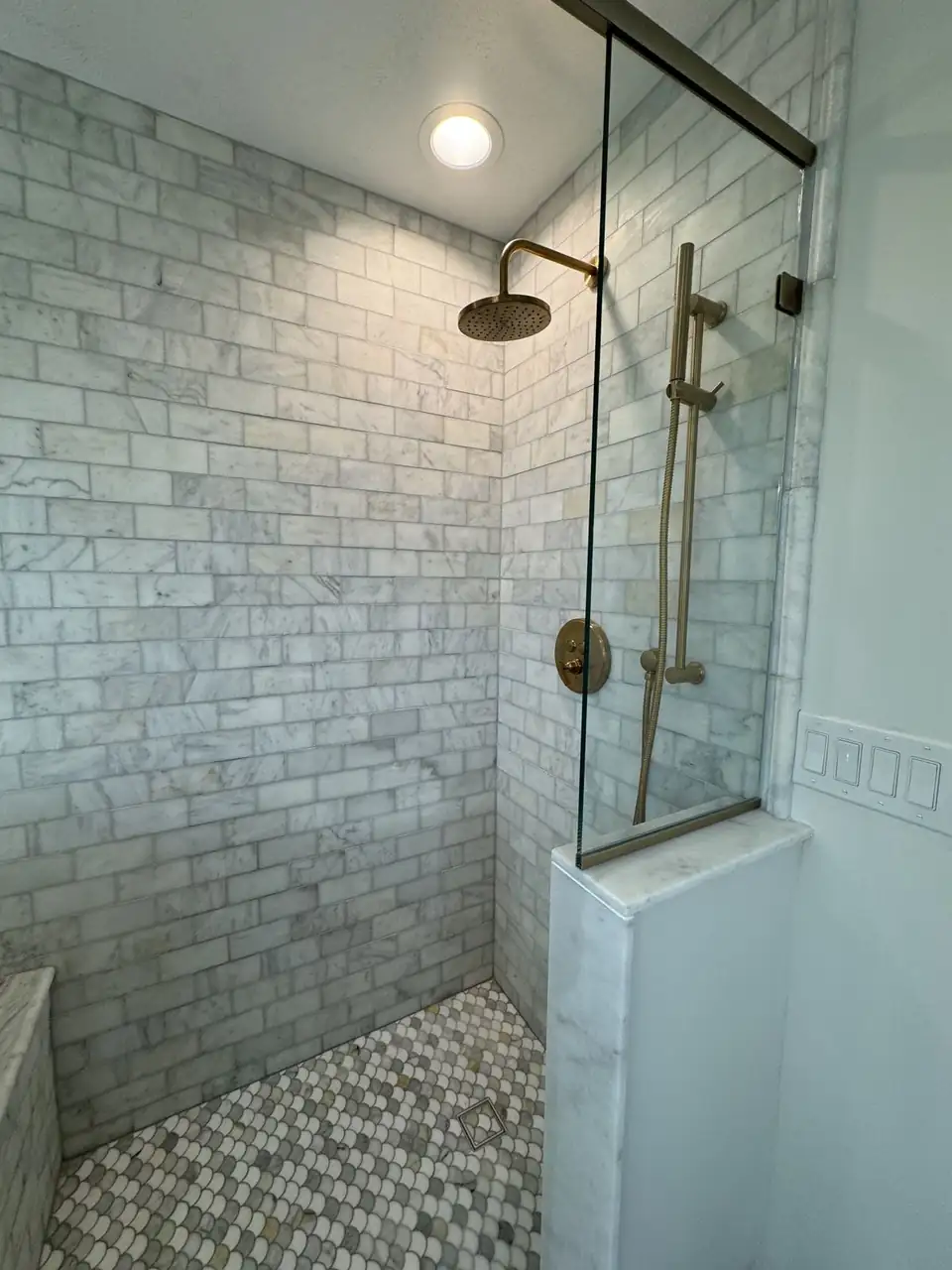 Tile installation in a walk-in shower: white marble subway tile walls, marble fish-scale mosaic floor, marble bench and brushed gold fixtures by Neptoon Tile Company